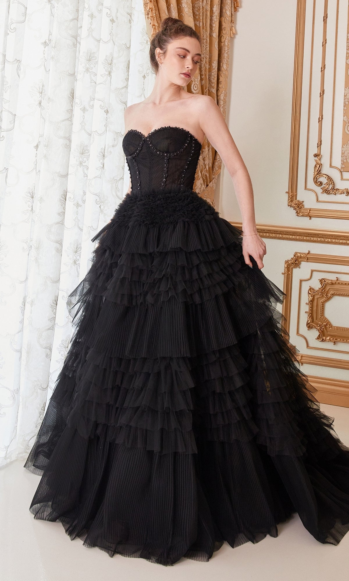 A woman in the Strapless Tiered Long Prom Ball Gown A1017, featuring a fitted bodice and layered skirt, stands in an ornate room with gold-trimmed walls and white curtains.